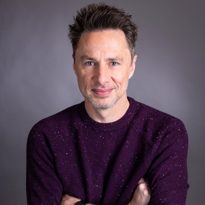 A man in a purple sweater and striped beige pants stands with his arms crossed.