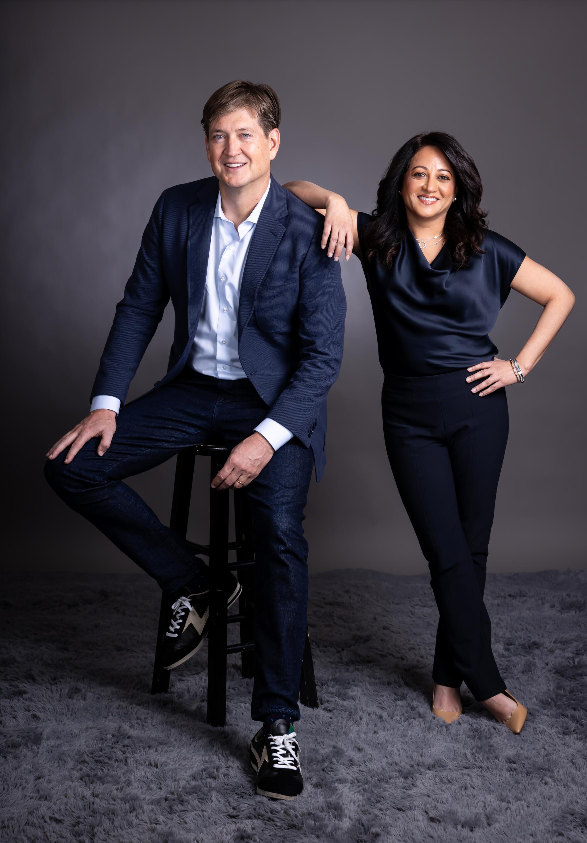 A man in a navy suit sits on a stool and a woman in a navy top and black slacks leans against his shoulder.