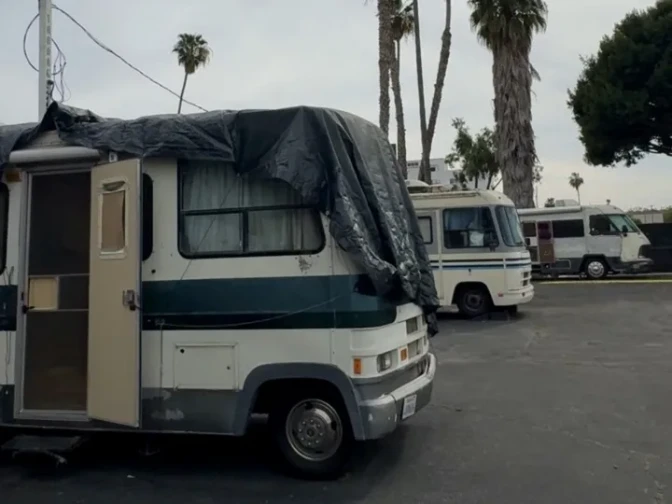 RVs are parked in a parking lot with some trees in the background. One of the RVs is covered with a tarp.