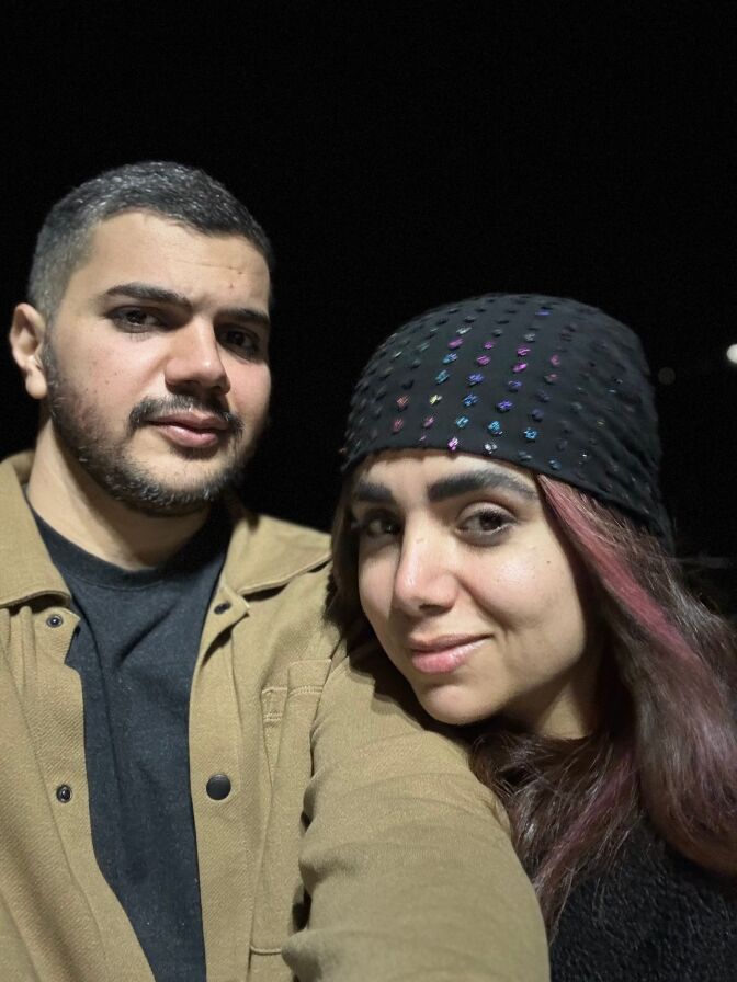 A couple take a selfie. He wears a brown jacket and black T-shirt. She wears a black bandanna.