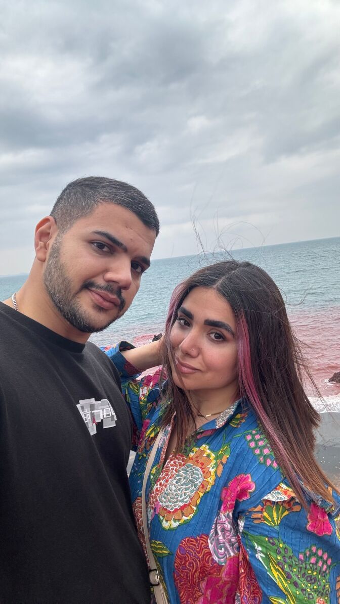 A couple stand in front of the ocean. The man wears a black T-shirt and the woman has pink streaks in her hair and wears a colorful top.