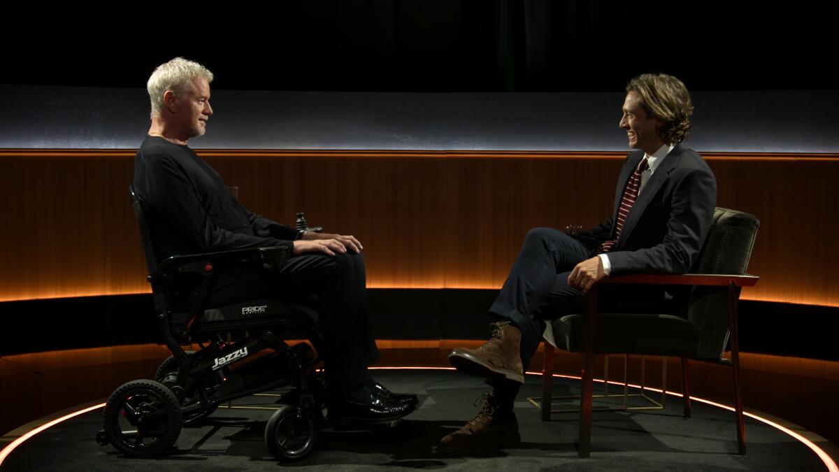 Two men sit across from each other in a dark room with wood paneling and back lighting.