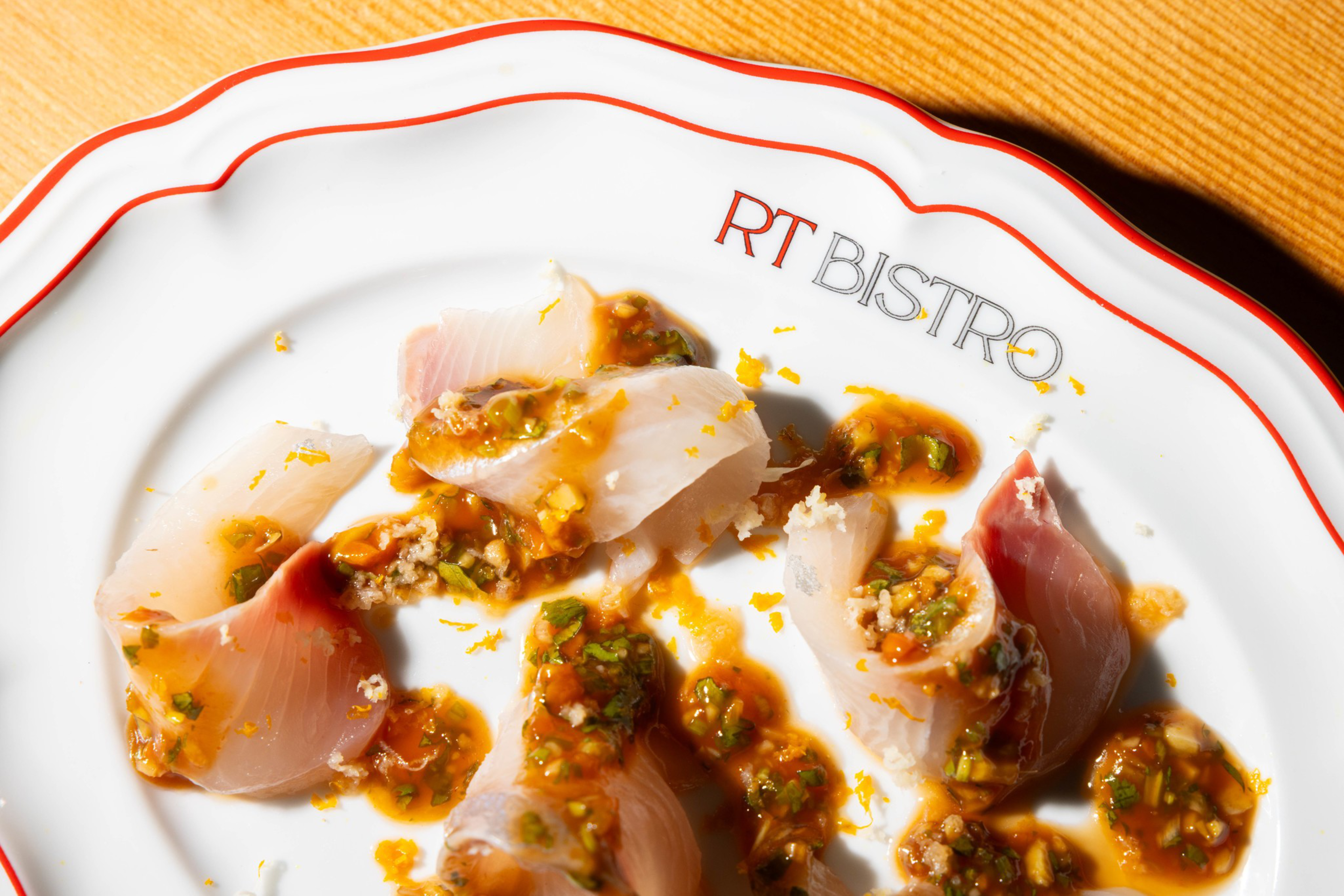 Thin slices of raw fish are arranged on a white plate with orange and green sauce drizzled over them, placed on a wooden surface.