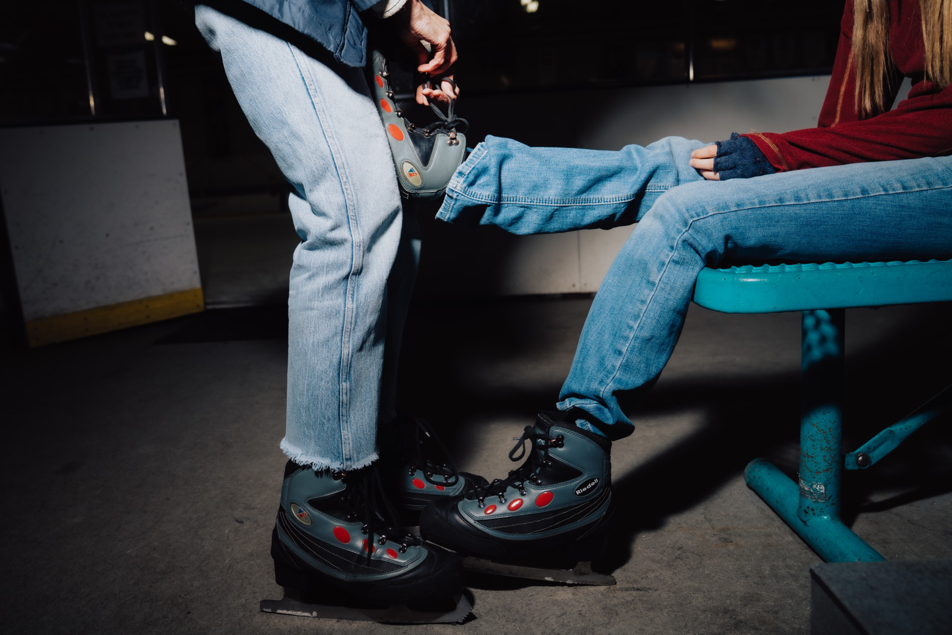 One person helps another put on ice skates while the second sits on a bench, both wearing jeans and black-and-gray skates with red accents.