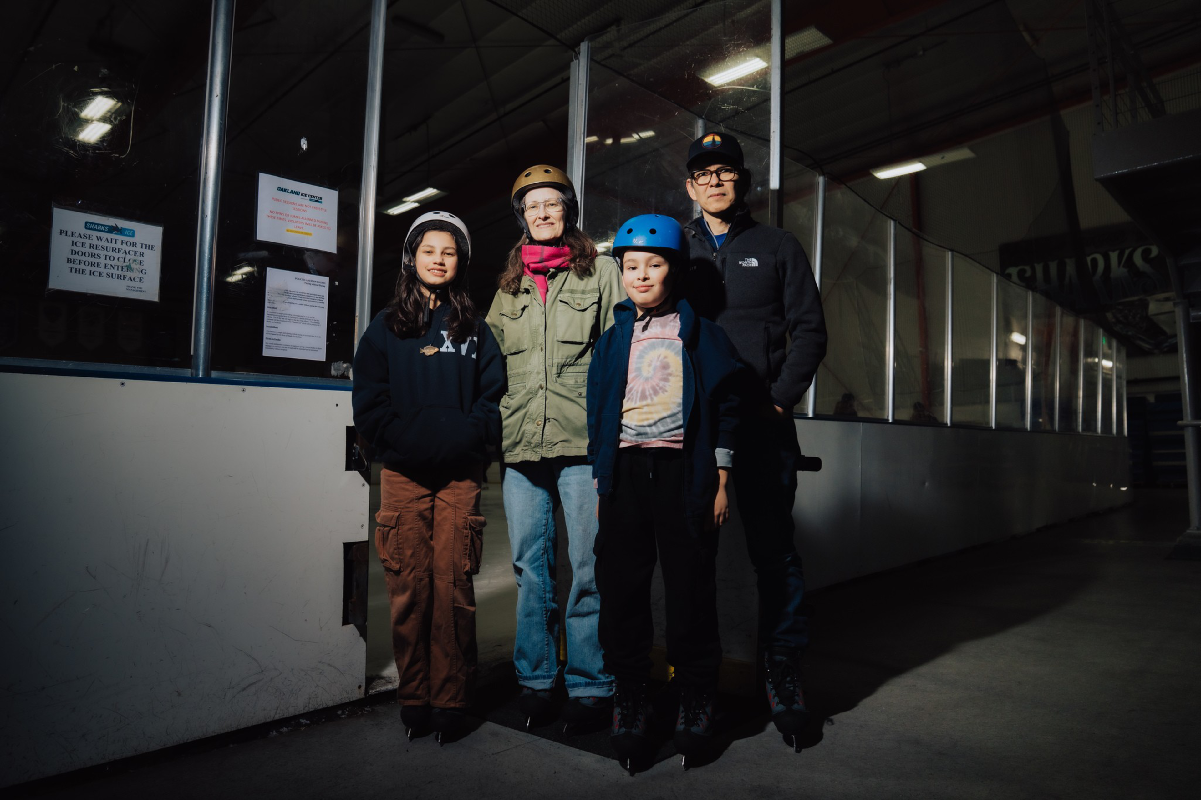 Four people wearing helmets and rollerblades stand on a rink floor, illuminated by a single light against a dark, empty rink background.