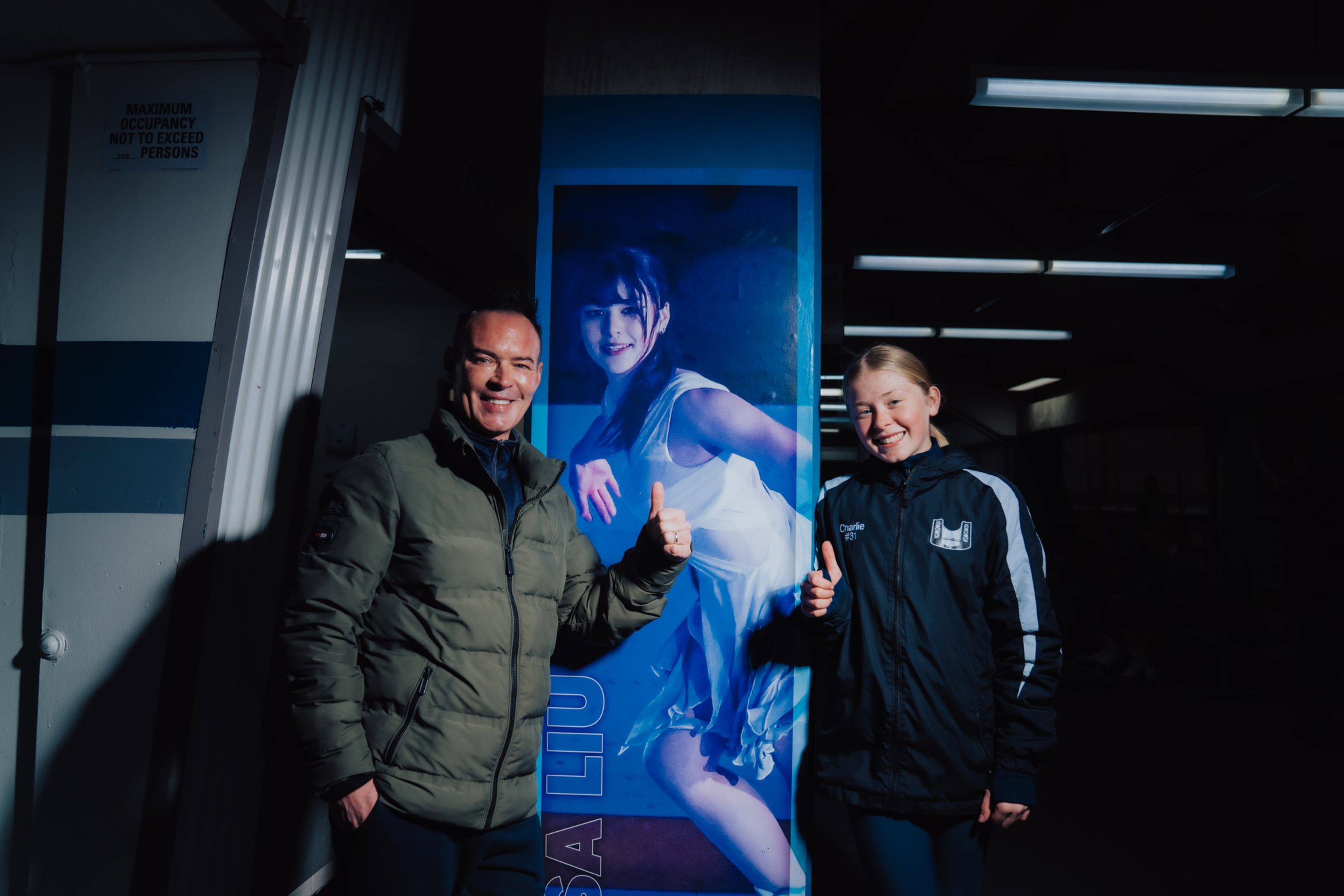 Two people smile and give thumbs up while standing on either side of a large poster of a female figure skater.