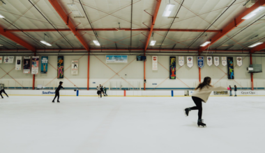 Several people ice skate inside a large indoor rink with banners hanging on the walls and a high ceiling supported by orange beams.