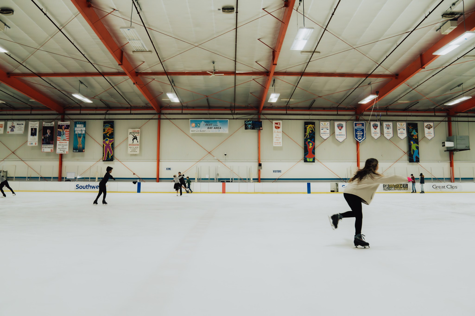 Several people ice skate inside a large indoor rink with banners hanging on the walls and a high ceiling supported by orange beams.
