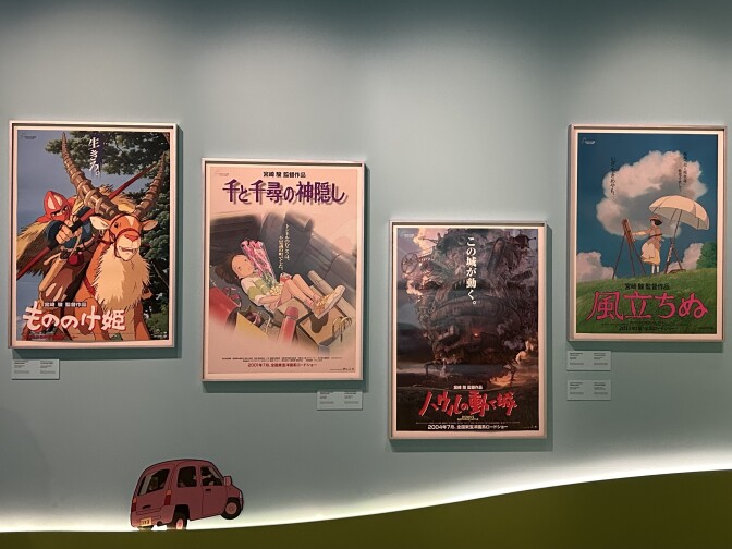 Four Japanese posters are hung on a light blue wall in silver frames from the movies Princess Mononoke, Spirited Away, Howl’s Moving Castle and The Wind Rises.