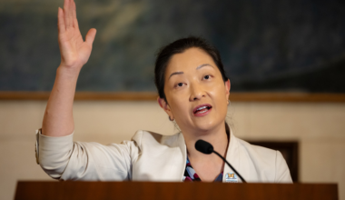 A woman in a light-colored blazer speaks behind a podium with one hand raised beside a microphone.