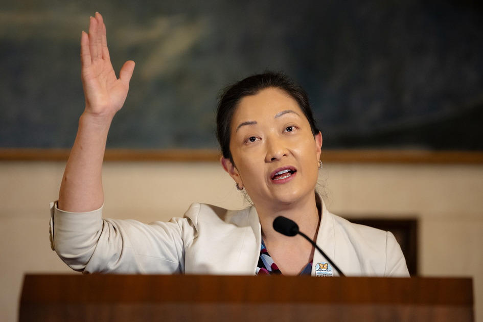 A woman in a light-colored blazer speaks behind a podium with one hand raised beside a microphone.