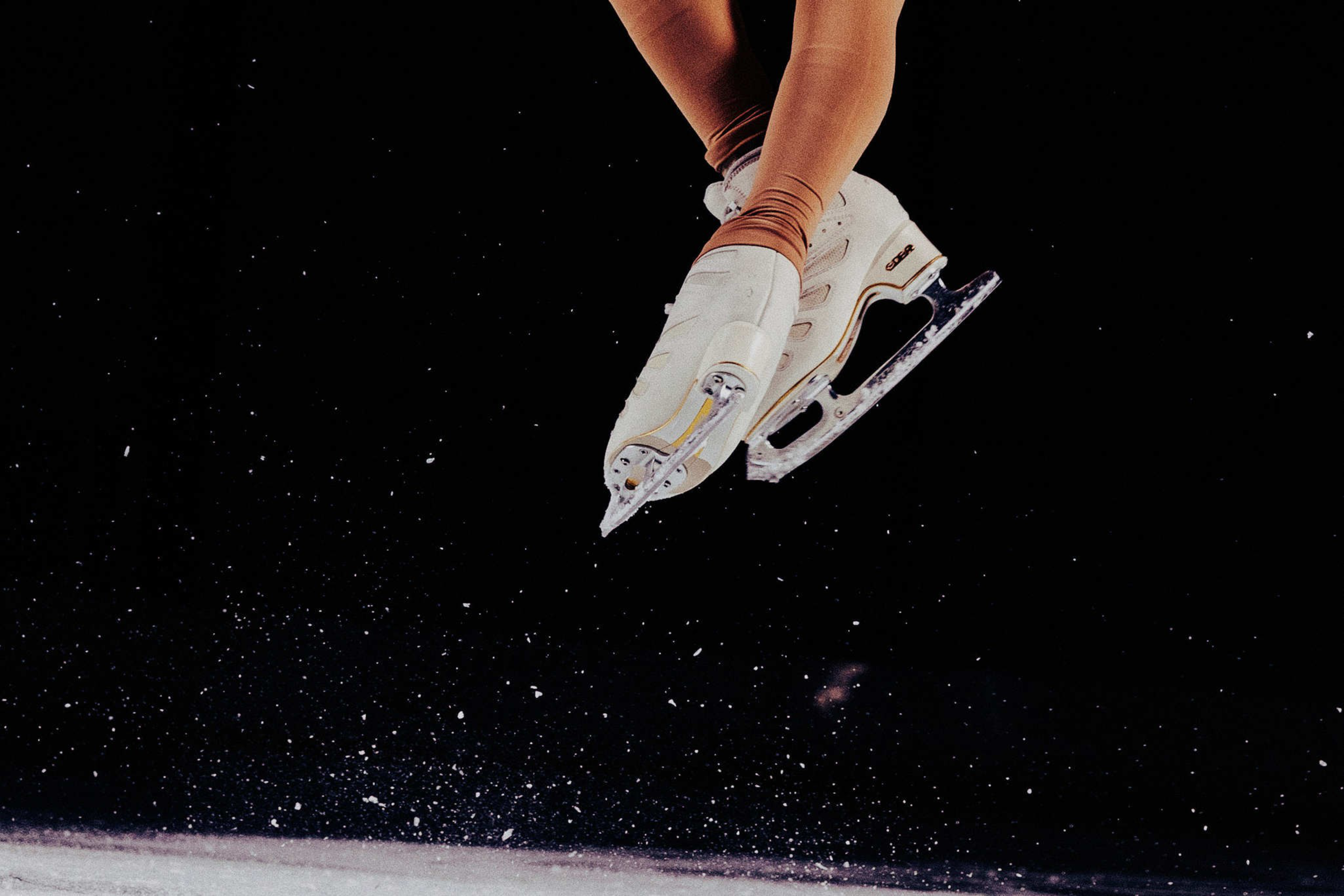 A pair of legs in brown tights wearing white ice skates are captured mid-air above an ice rink with ice particles scattered around.