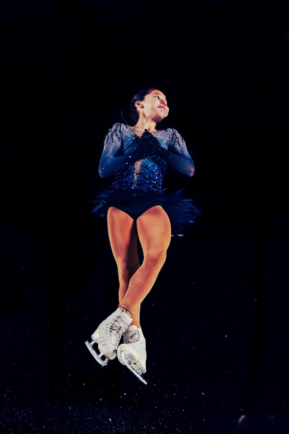 A figure skater in a dark blue dress and white skates leans against a wall in a concrete hallway near closed double doors labeled "Referees Room 6."