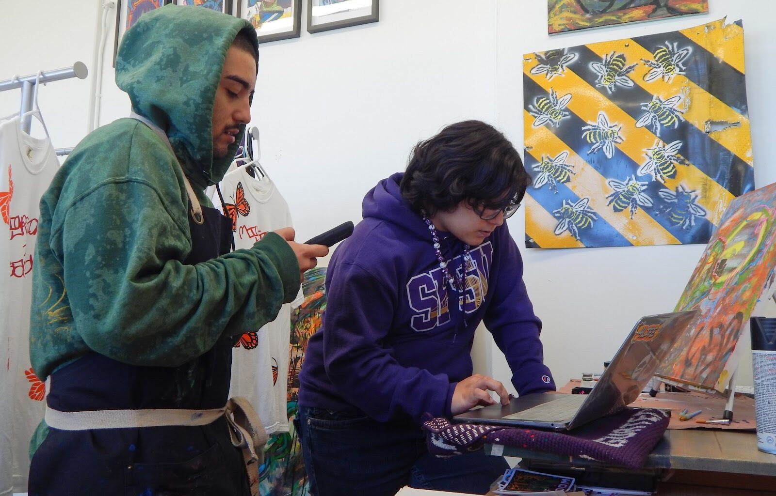 Two people work in an art studio; one uses a laptop while the other looks at a phone. Painted shirts and bee-themed artwork are visible in the background.