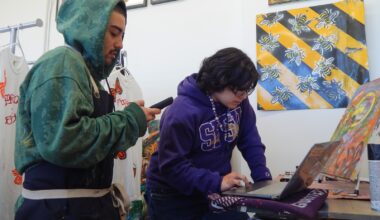 Two people work in an art studio; one uses a laptop while the other looks at a phone. Painted shirts and bee-themed artwork are visible in the background.