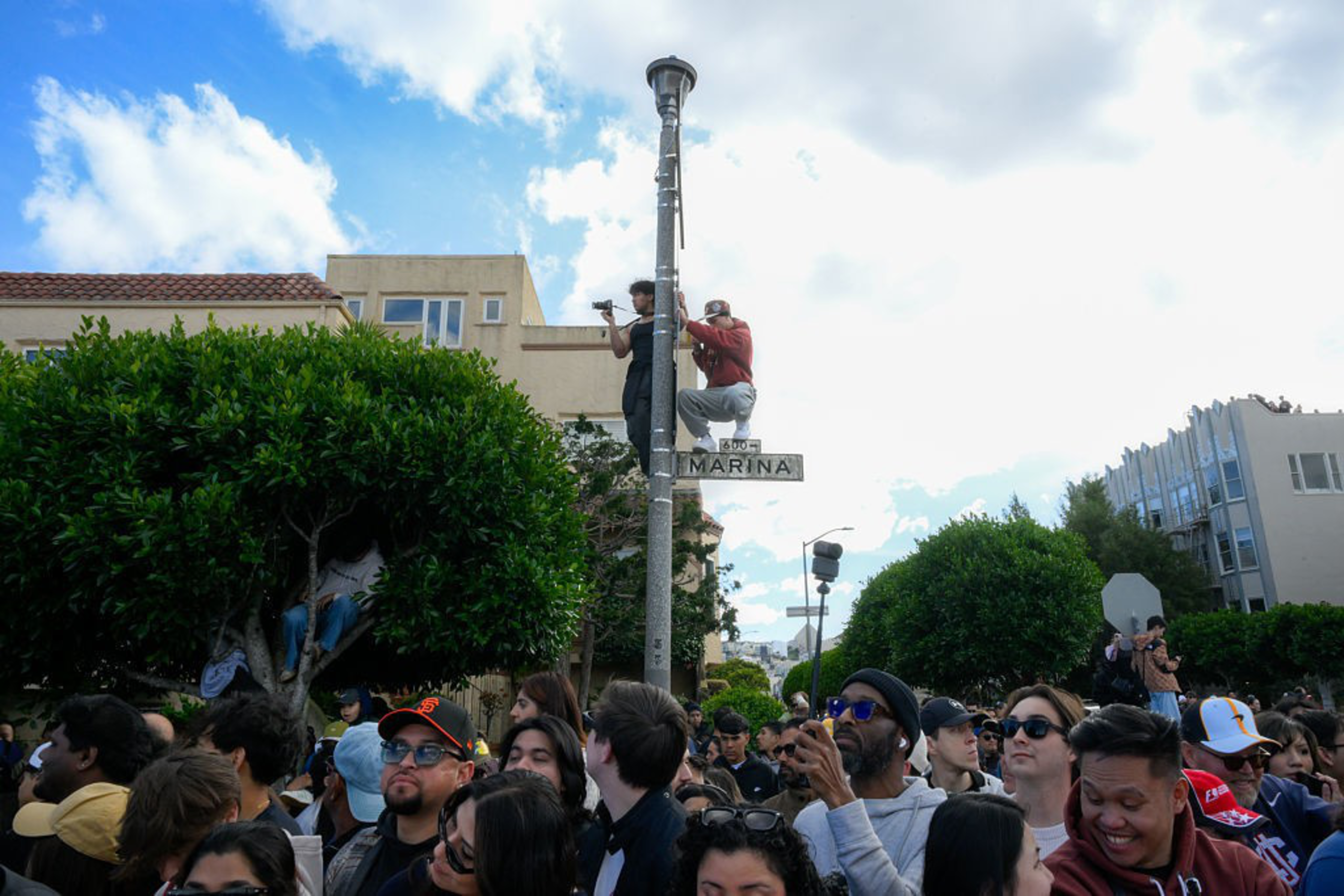 Two people are perched on a streetlamp marked “Marina” above a dense crowd, with some individuals also sitting in trees nearby.