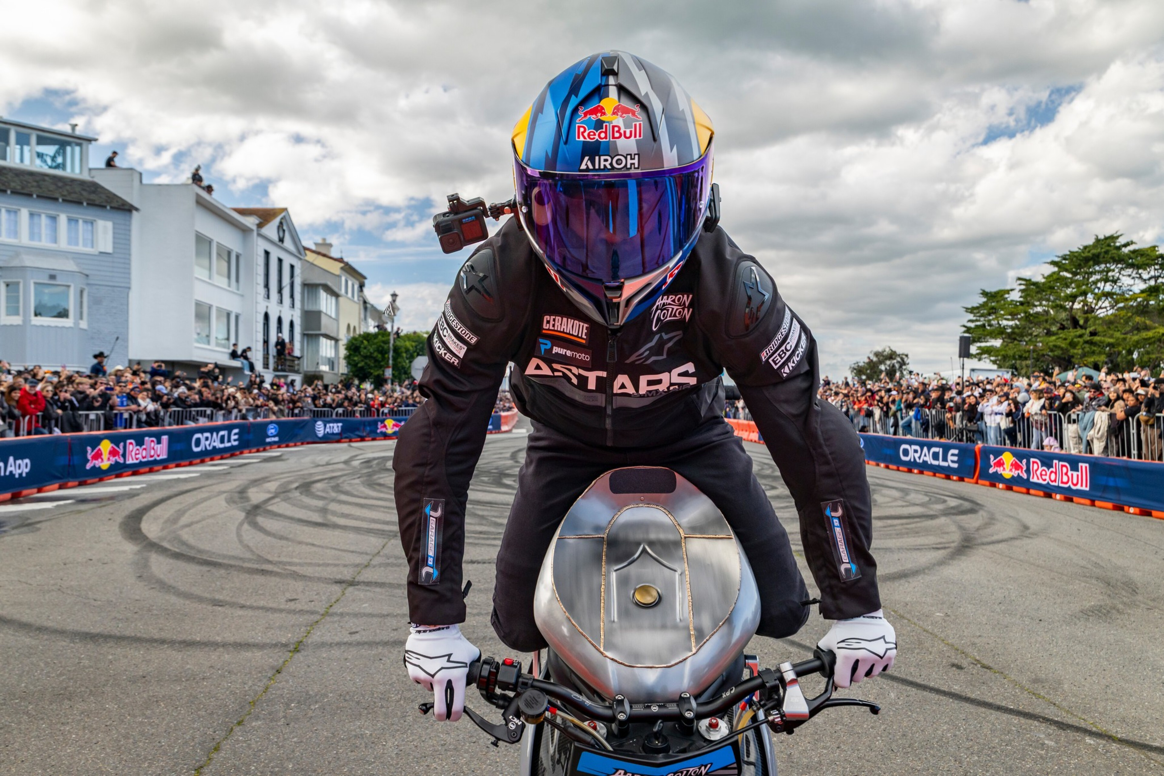 A motorcyclist wearing a helmet and racing suit performs a stunt on a metal motorcycle amid tire marks and a crowd behind barriers.