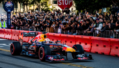 A Red Bull Formula 1 car races on a city street lined with red barriers and a crowd of spectators taking photos behind metal fences.