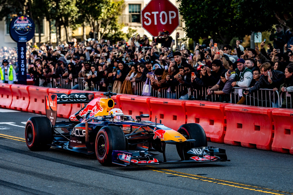 A Red Bull Formula 1 car races on a city street lined with red barriers and a crowd of spectators taking photos behind metal fences.