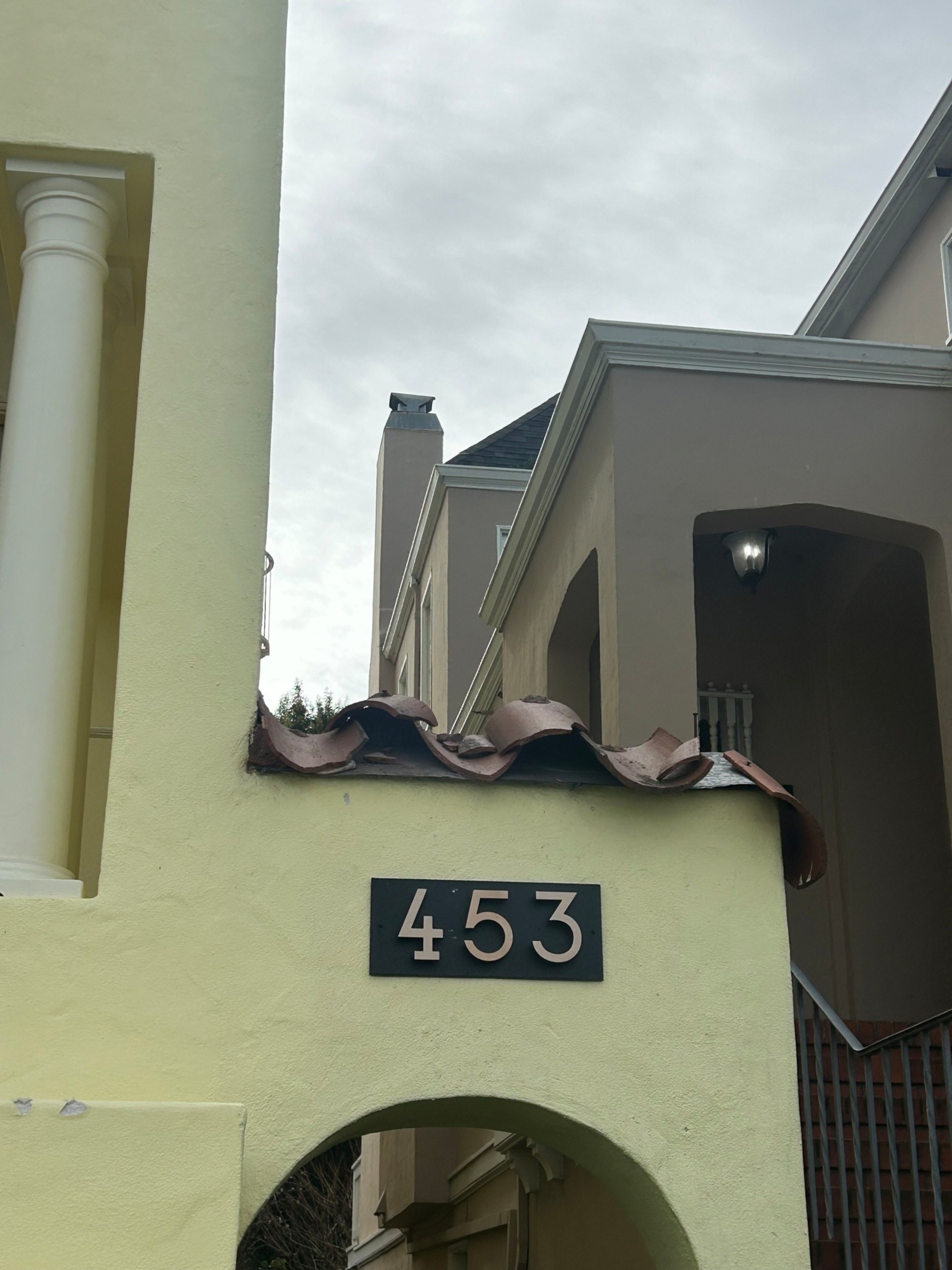 A light yellow wall with a black address plaque showing the number 453, partially damaged roof tiles, and beige buildings with stairs and a lit porch light.