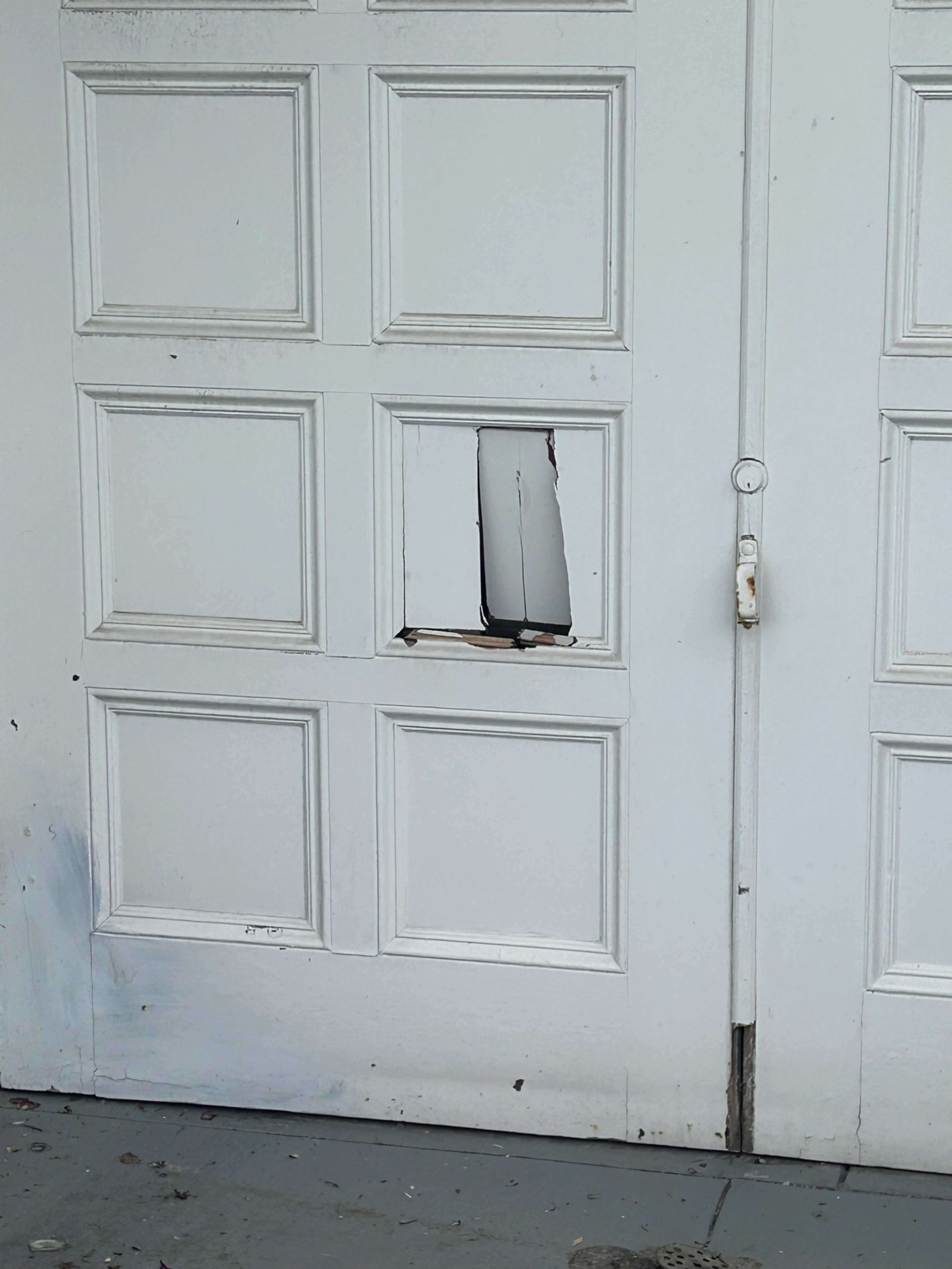 A white paneled door with a rectangular hole in one panel and chipped paint around the hole and edges, revealing the interior beneath.