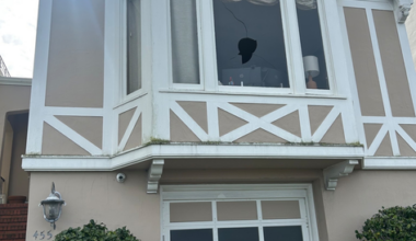 A two-story beige house with white trim has a garage door below a bay window, a broken window pane, bushes on either side, and stairs leading up on the left.