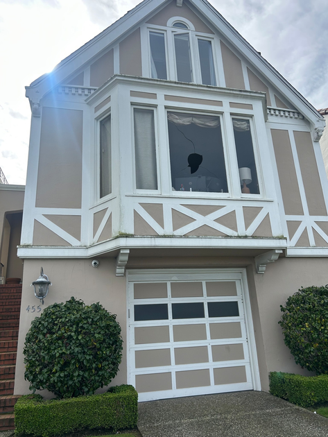 A two-story beige house with white trim has a garage door below a bay window, a broken window pane, bushes on either side, and stairs leading up on the left.