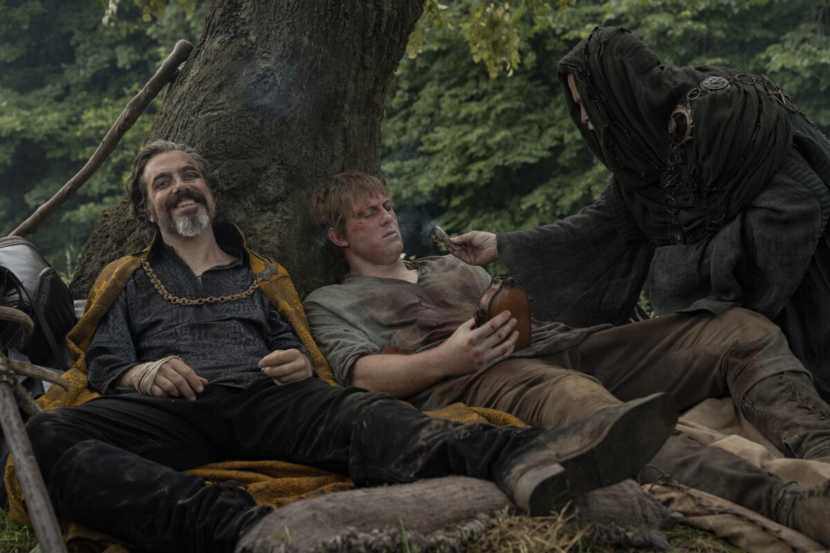 two injured men leaning against a tree while another examines them