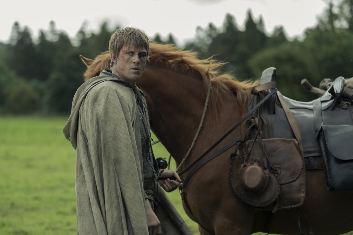 a man in cloak standing next to a horse and holding its reins