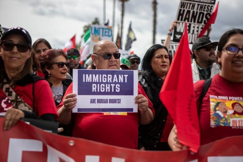 A person in sunglasses and a red shirt holds up a sign that reads, “Immigrant Rights are Human Rights!,” while standing in a crowd holding more signs.