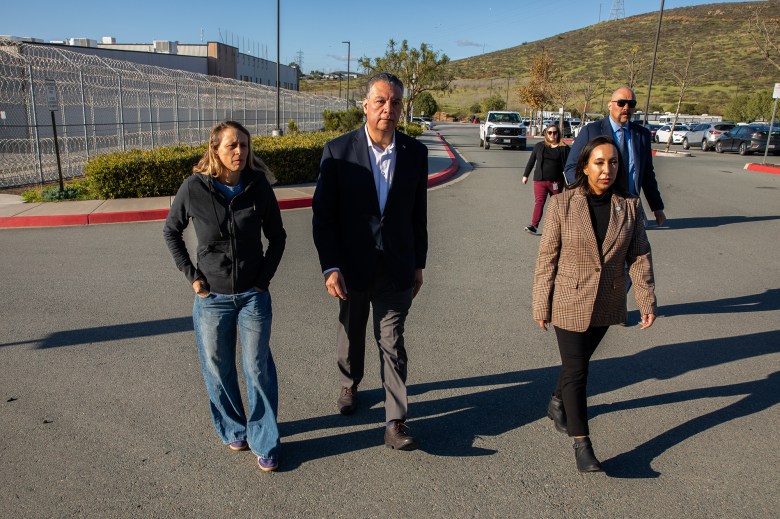 A group of officials and staff walk down a roadway beside a high chain-link fence topped with razor wire outside a large facility, with parked cars and low hills in the background under bright afternoon sun.