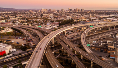 Curving elevated highways intersect above a busy urban area with numerous cars, leading toward a distant city skyline under a warm, pink-tinted sky.