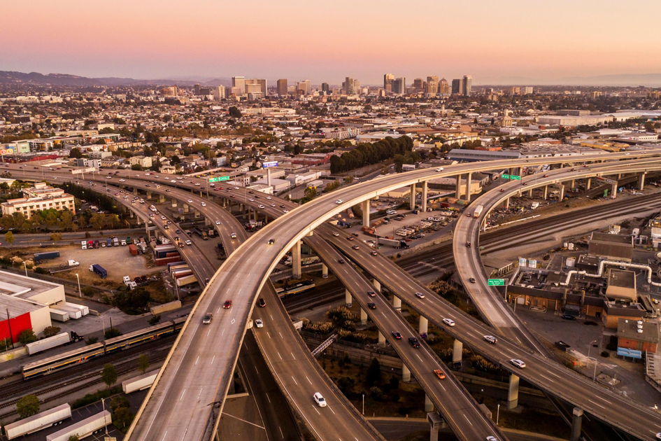Curving elevated highways intersect above a busy urban area with numerous cars, leading toward a distant city skyline under a warm, pink-tinted sky.