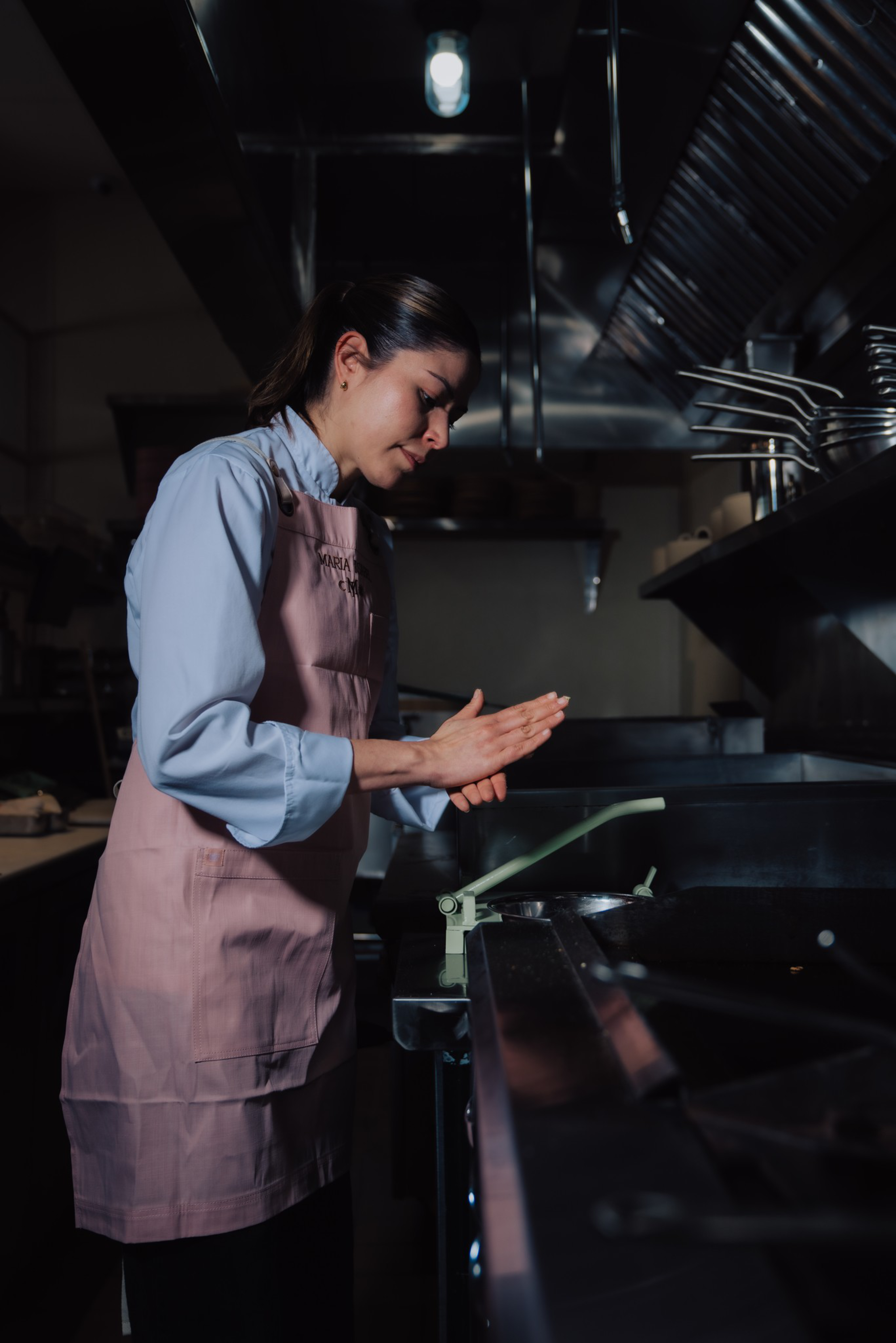 A woman wearing a pink apron operates a green kitchen press, preparing food in a dimly lit commercial kitchen.
