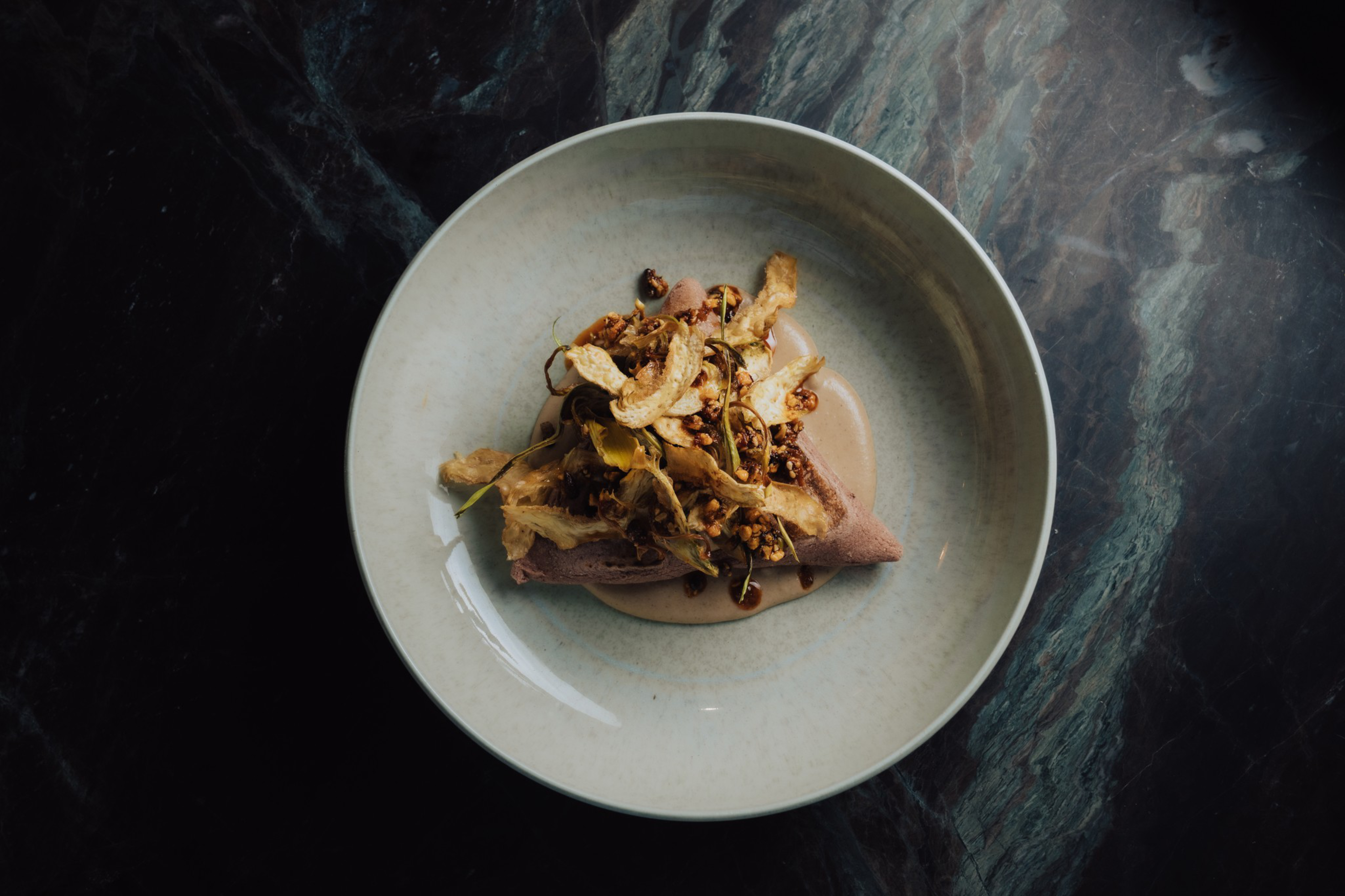 A folded crepe topped with granola, crispy vegetable chips, and drizzled with chocolate sauce on a beige plate over a dark marble surface.