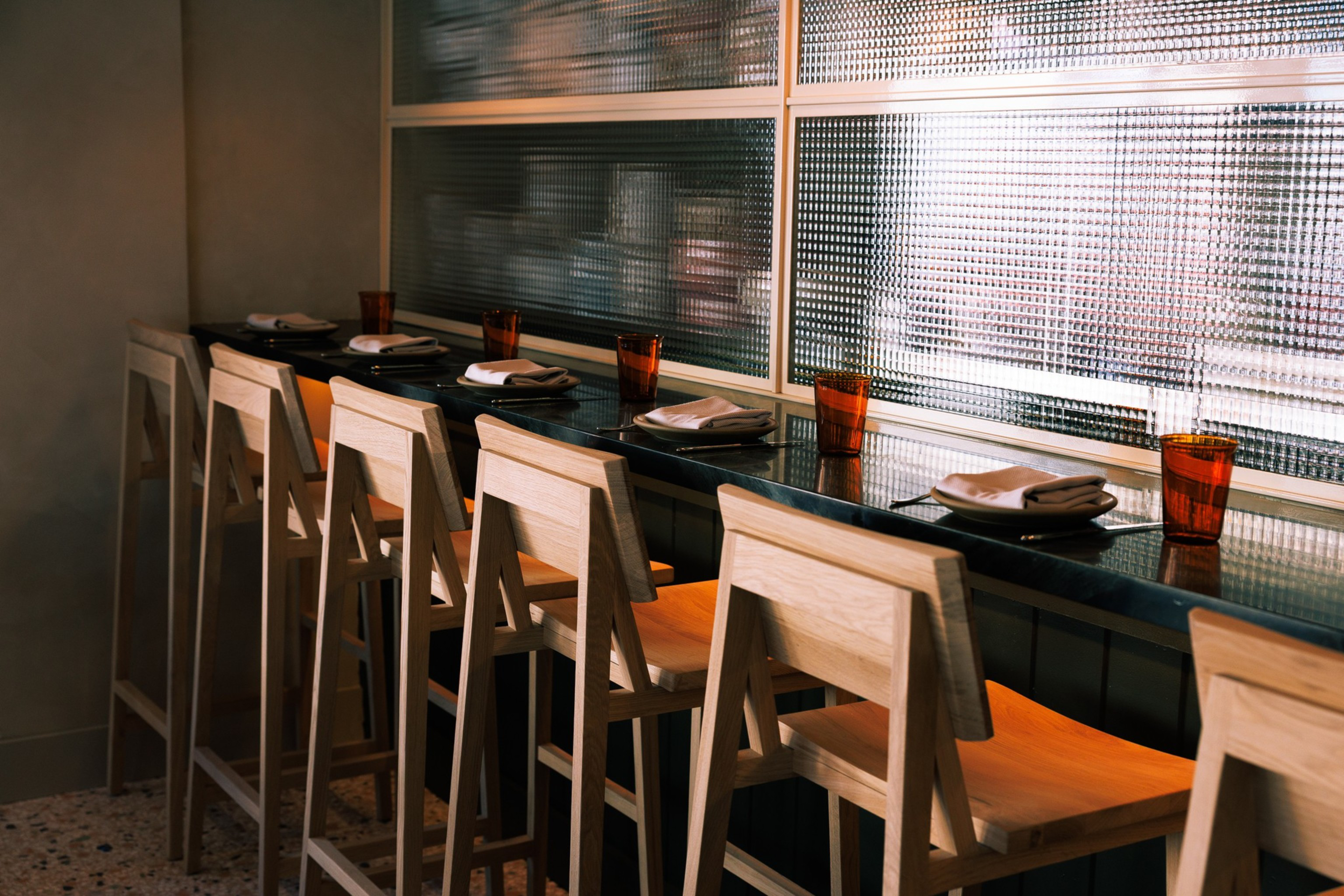 Seven wooden bar stools are lined up at a dark counter with orange-tinted glasses and folded napkins on white plates in front of each seat.