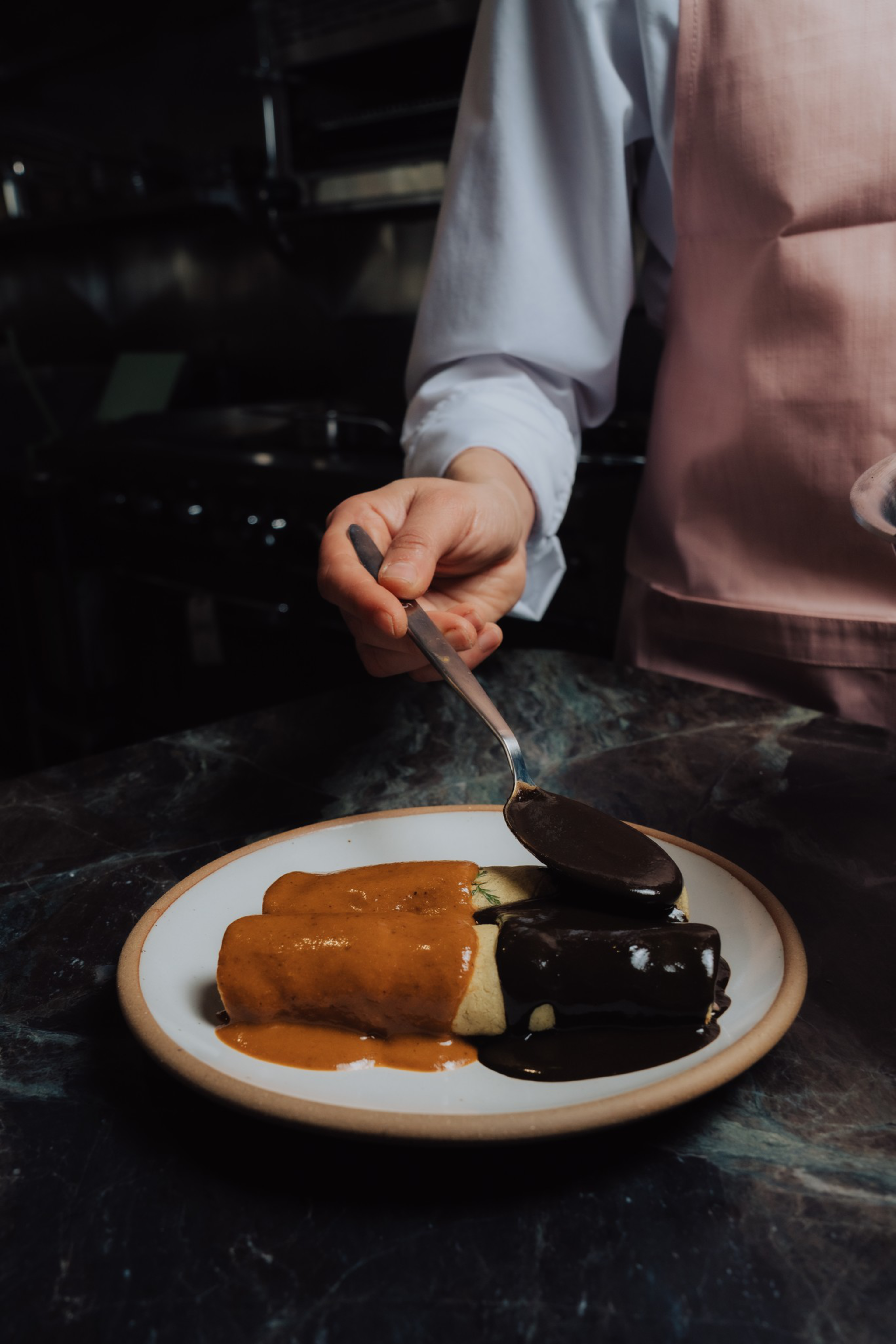 A person in a pink apron is spooning dark sauce over two rolled tortillas, one covered in brown sauce and the other in dark sauce, on a white plate.