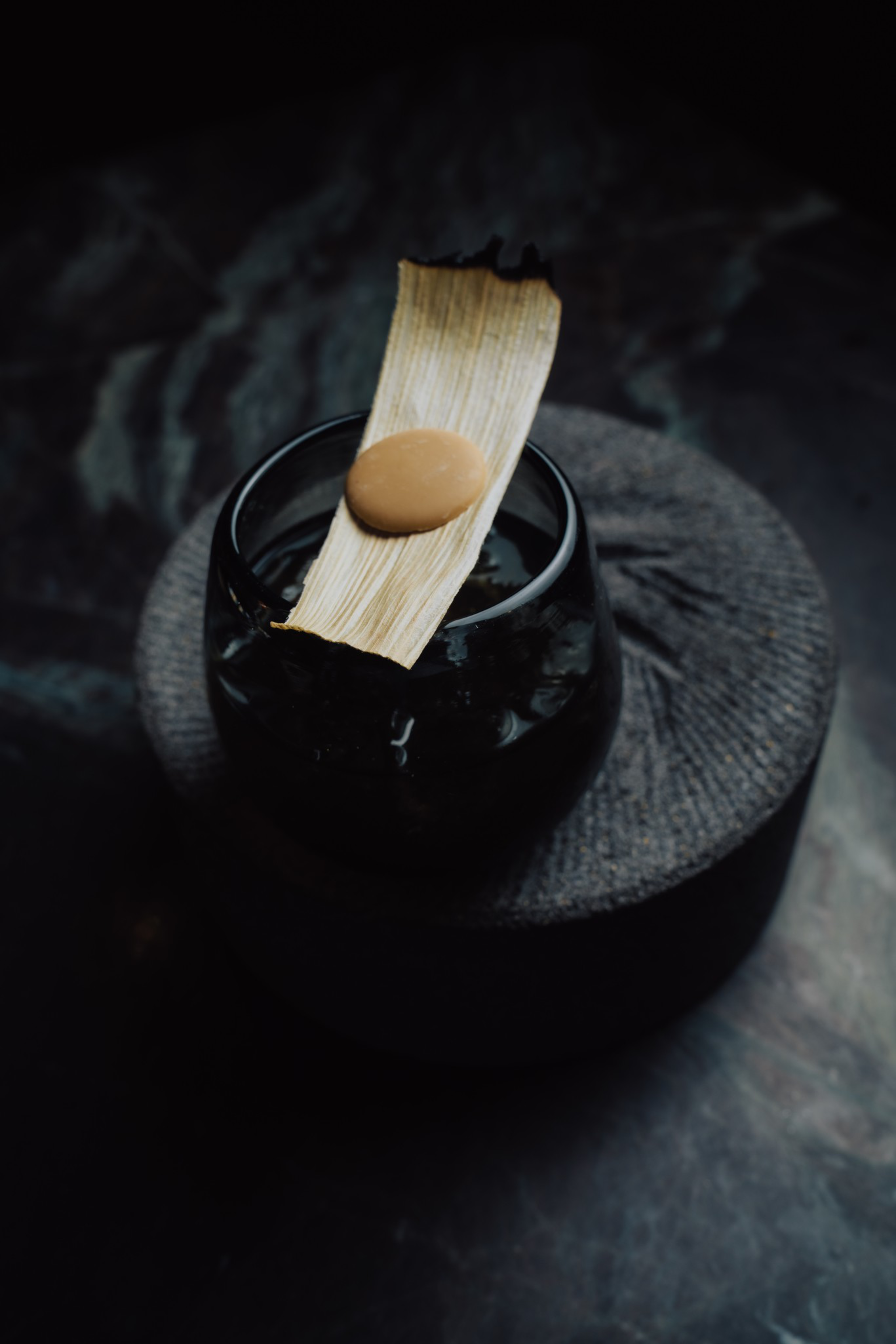 A dark glass with ice sits on a textured stone surface, topped with a dried leaf holding a small round beige object.