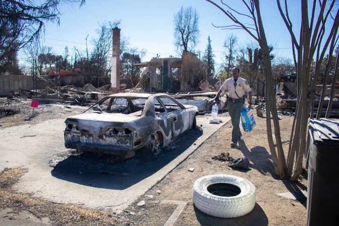 A Black man wearing a tan uniform with a badge is carrying a large bag of cat food in one hand and a gallon of water in the other through the remains of a burned-out property and home in Altadena.