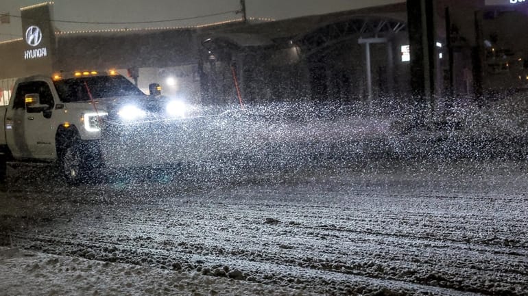 A snow plow’s headlights cut through the darkness, illuminating the...