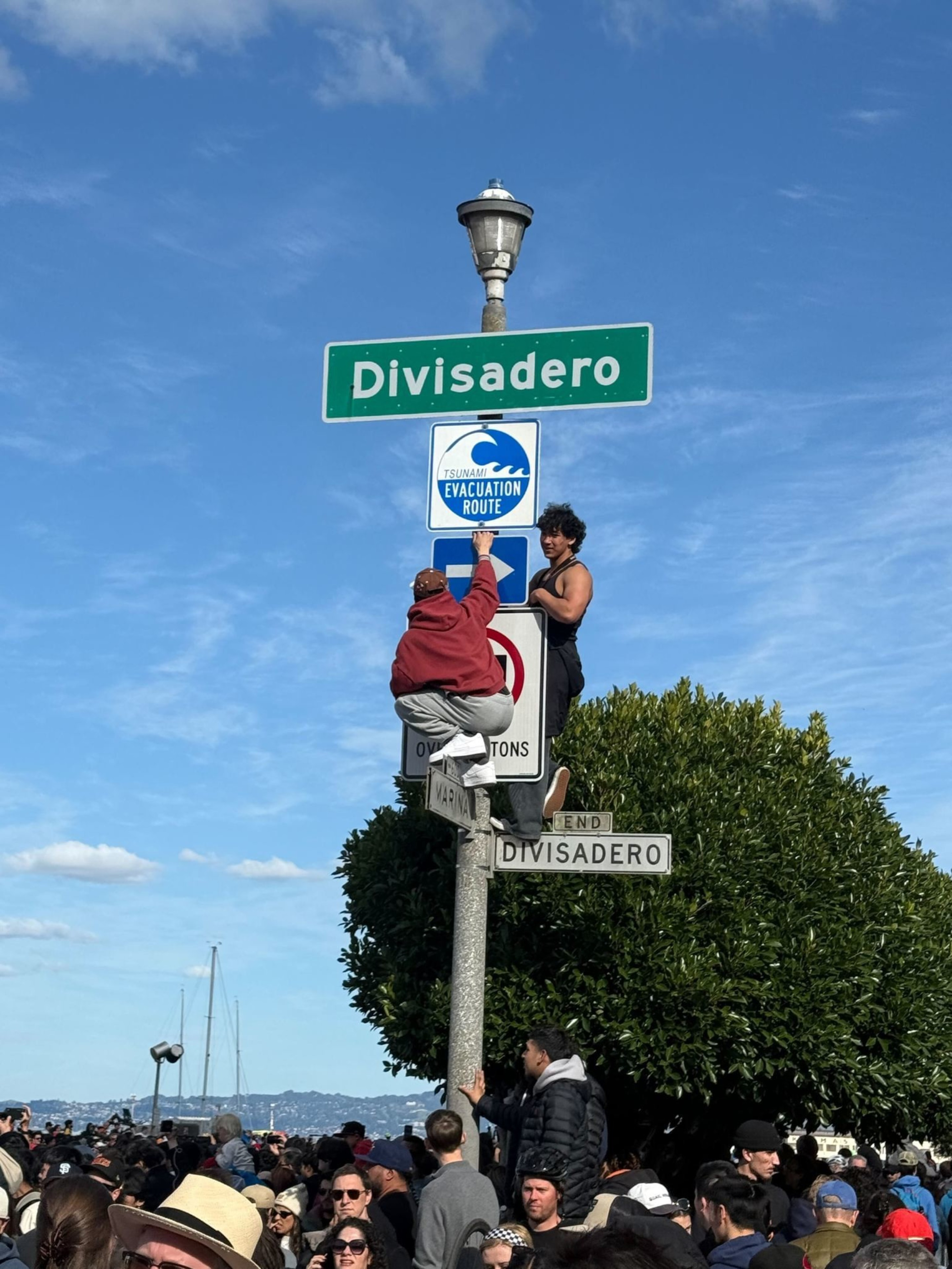 Two people are climbing a street sign pole labeled “Divisadero” and “END DIVISADERO,” above a large crowd gathered on a sunny day by the water.