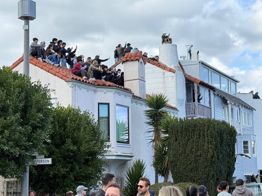 Two people are climbing a street sign pole labeled “Divisadero” and “END DIVISADERO,” above a large crowd gathered on a sunny day by the water.