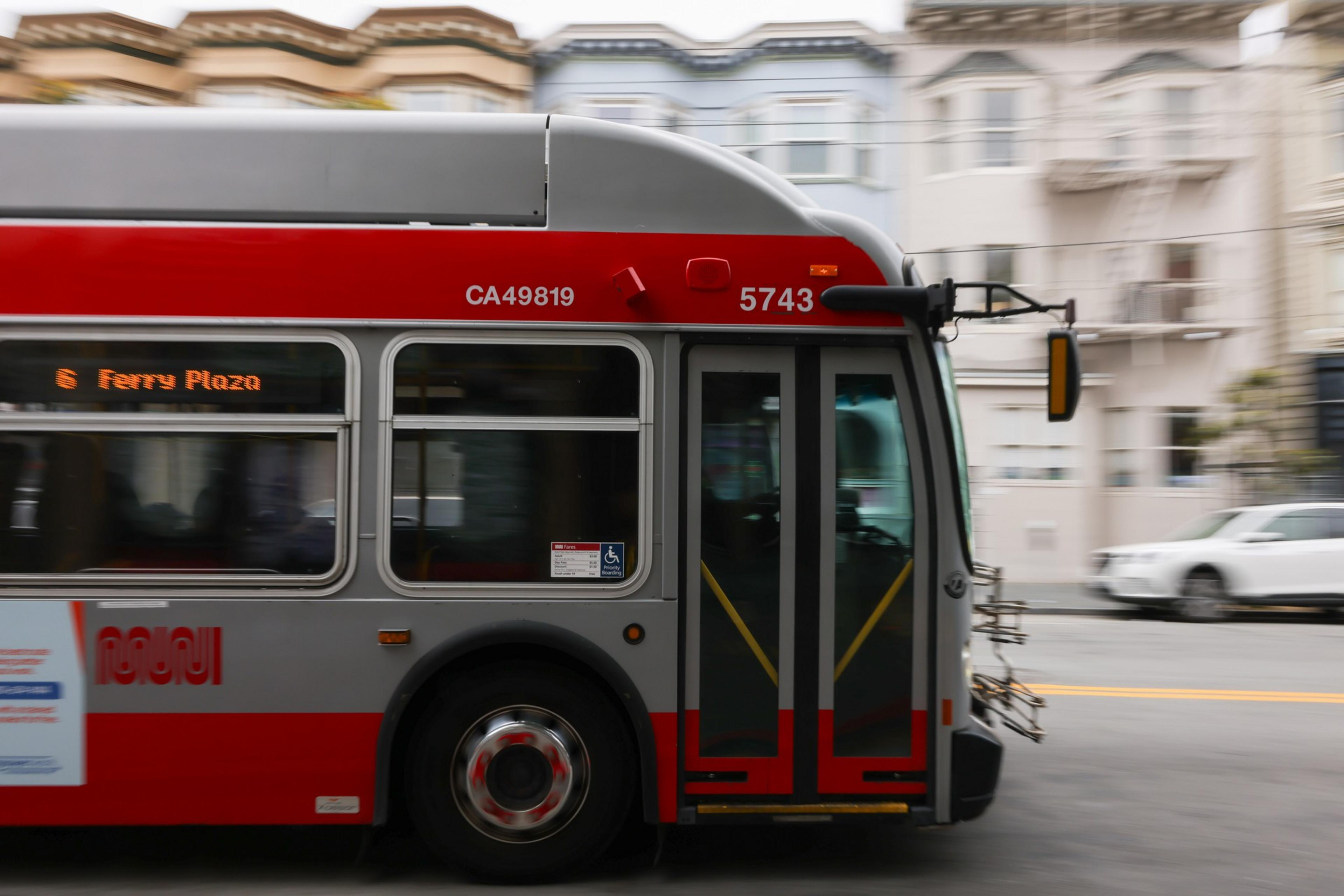 A red and gray city bus marked 5743 is in motion on a street with blurred buildings and cars in the background, displaying “Ferry Plaza” as its destination.
