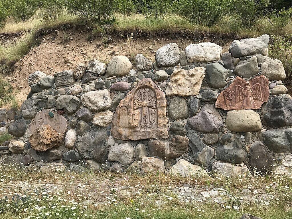 A photo of khackars, engraved stone, embedded into a rock wall on a grassy hillside with white wildflowers.