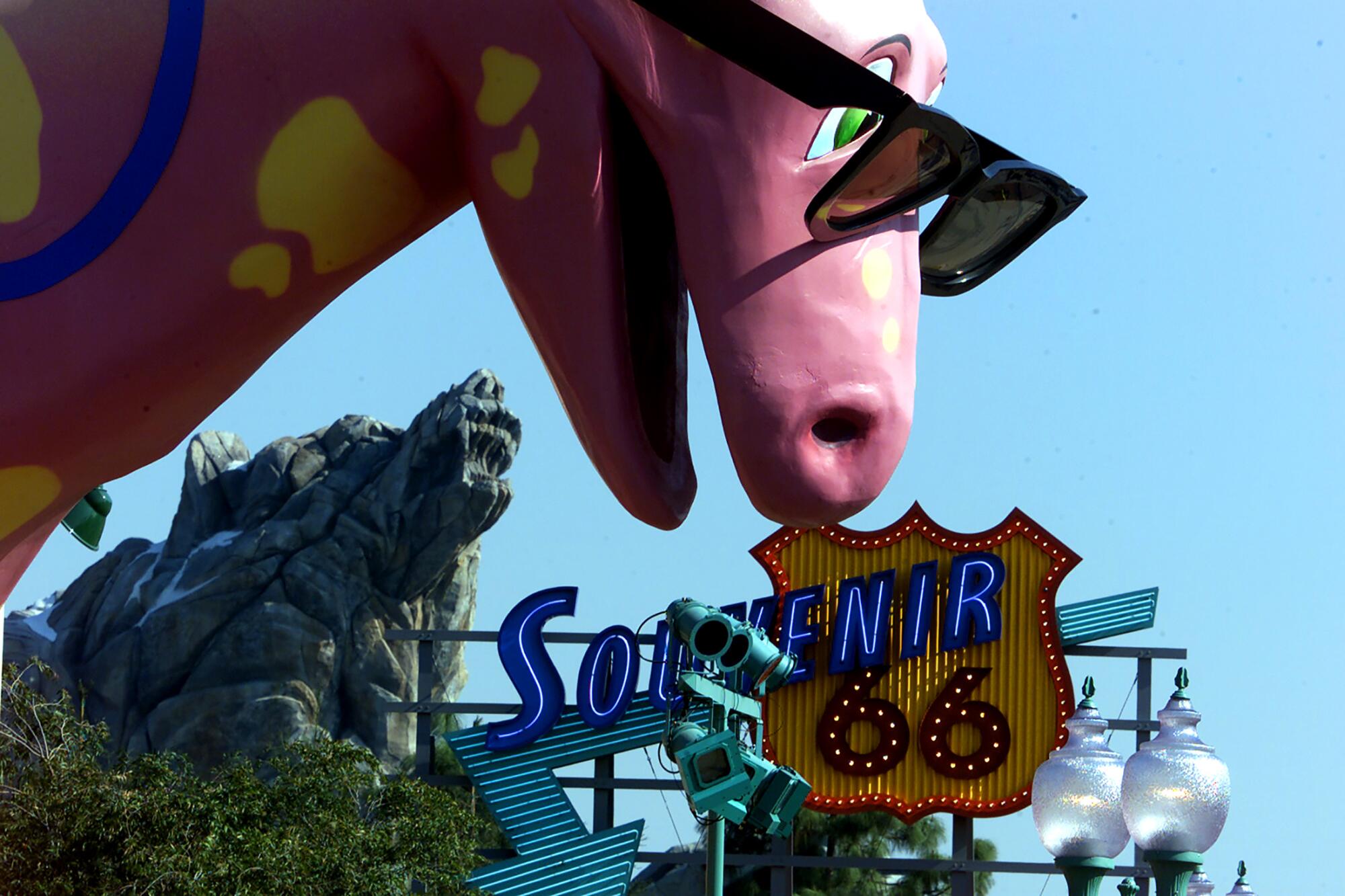 A pink dinosaur in sunglasses in a theme park, with a Route 66-themed shop in the background.