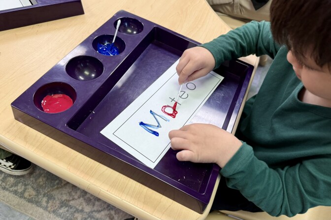 A student with light skin tone and brown hair traces a lower-case letter a in red paint with a q-tip. The paper he is painting has Mateo printed in black. 
