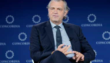 A man in a navy suit and dotted tie sits with legs crossed, gesturing with his hands, against a blue backdrop branded with "CONCORDIA" logos.