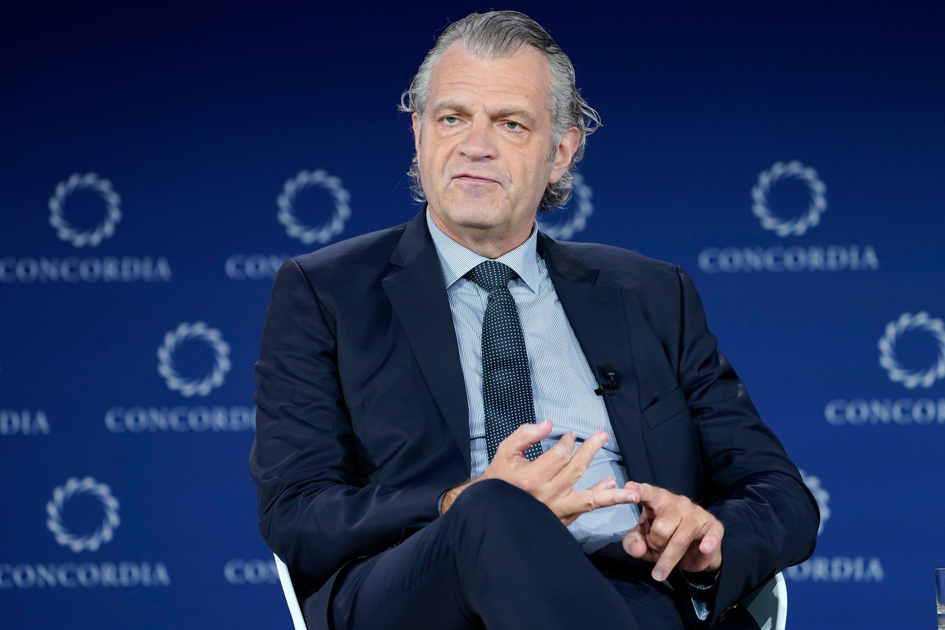 A man in a navy suit and dotted tie sits with legs crossed, gesturing with his hands, against a blue backdrop branded with "CONCORDIA" logos.