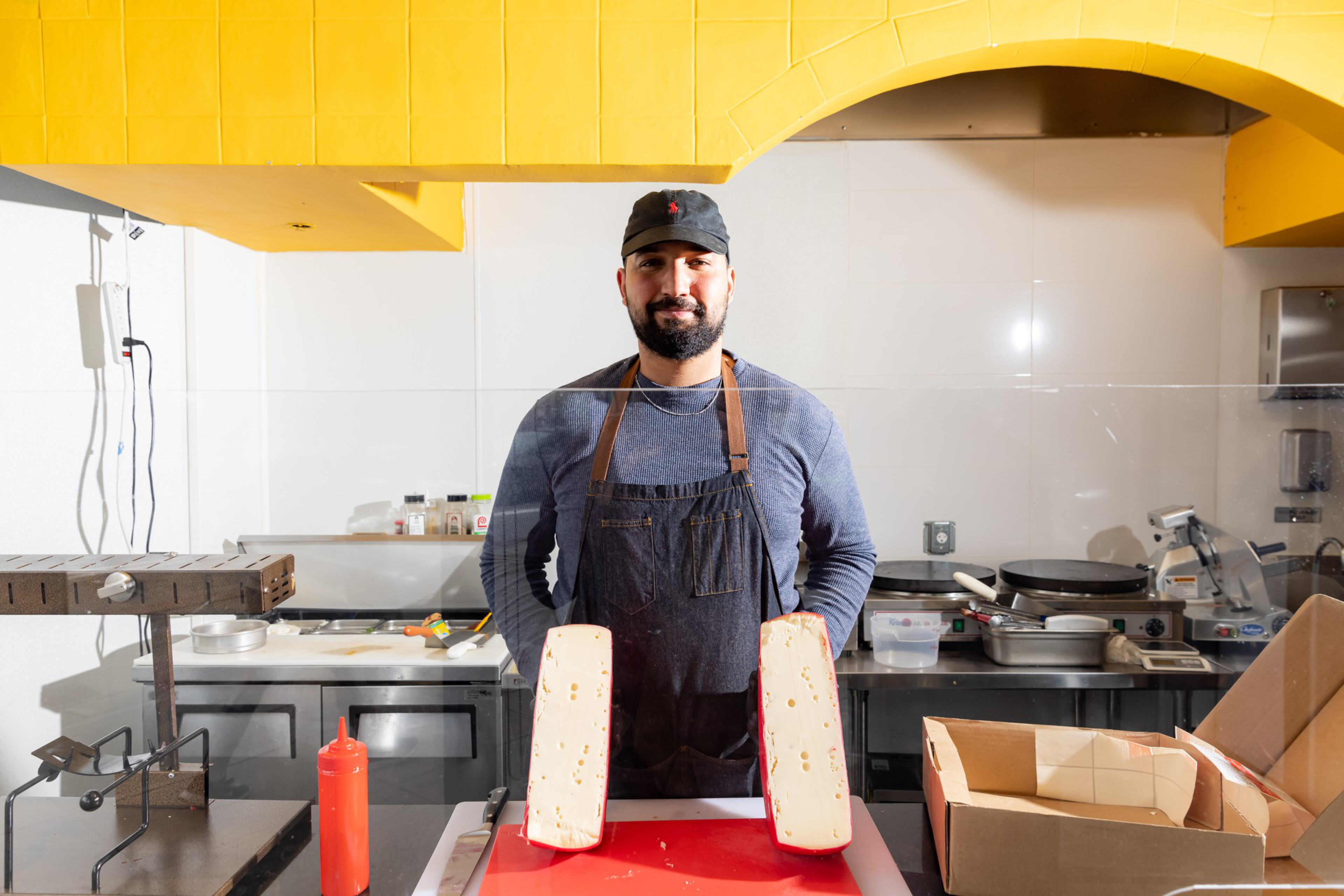 A man wearing a black cap, blue shirt, and black apron stands behind a counter with two large blocks of cheese cut in half.