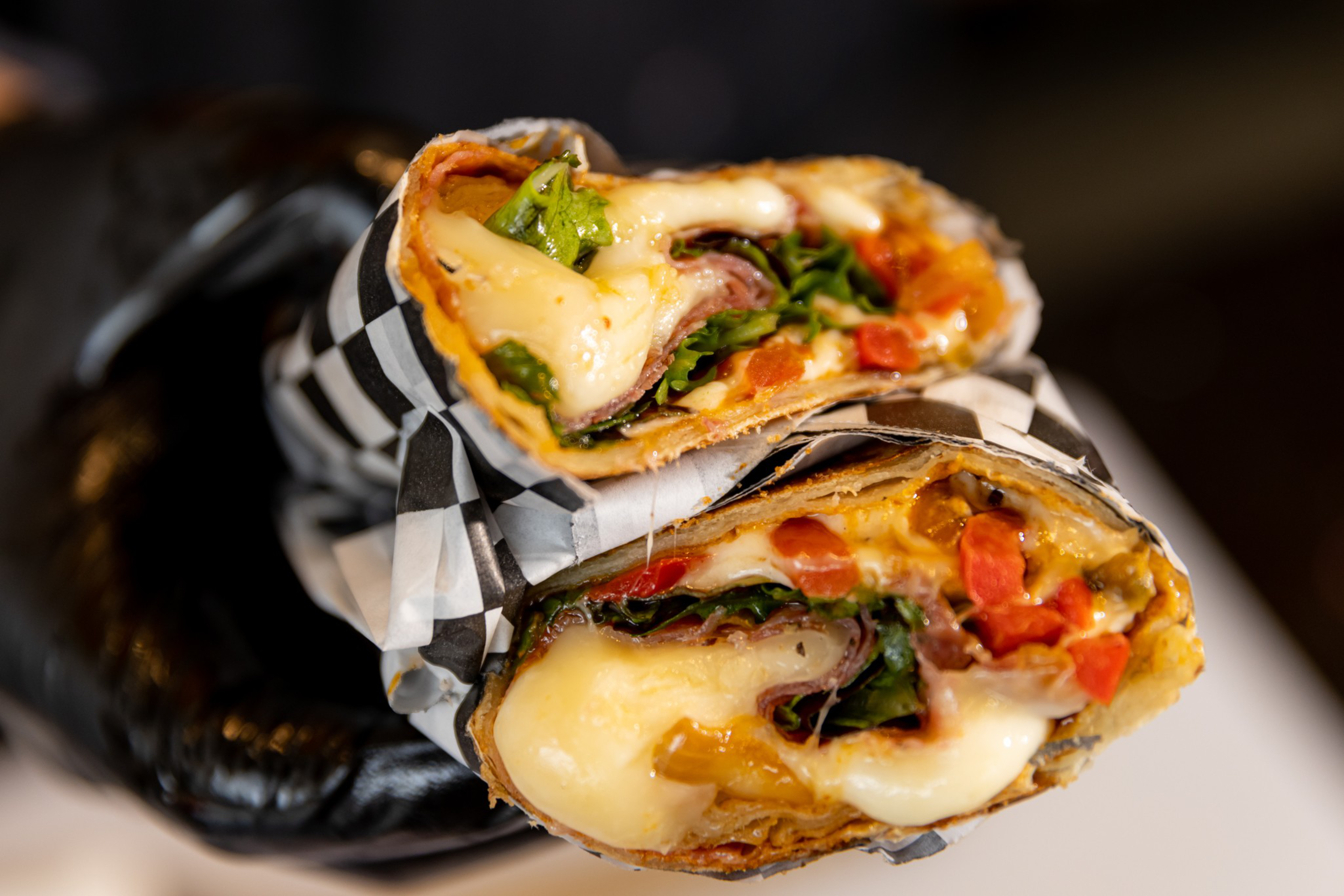 A grilled wrap cut in half reveals melted cheese, leafy greens, and diced red and yellow peppers, held by a gloved hand.